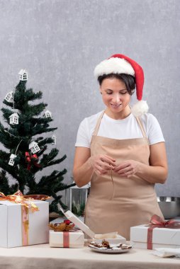 Mutfak önlüğü ve Noel Baba şapkası takan bir kadın Noel ağacı ve hediye kutularının yanında. Dikey çerçeve.
