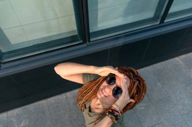 Siyah gözlüklü kırmızı rastalı güzel bir kız gökyüzüne bakıp gülümsüyor. Sokakta ateş ediyorlar. Mutlu kadın.