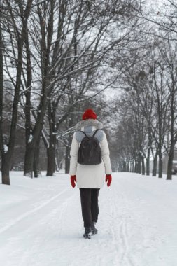 Kırmızı şapkalı ve eldivenli kadın parkın karla kaplı geçidi boyunca yürüyor. Karlı hava. Arka plan.