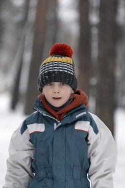 Ağaçların arkasındaki kış ormanındaki sevimli çocuk portresi. Örgü şapkalı ve kışlık ceketli çocuk doğrudan kameraya bakıyor. Dikey çerçeve.