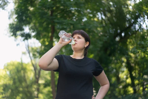 Young woman drinks water from the bottle after jogging in the spring park. Drinking regime during sports. Body positivity.