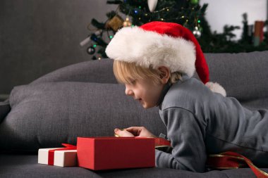 Noel Baba şapkalı çocuk, yılbaşı hediyesini Noel ağacının arka planında koltukta yatarken görüyor. Noel hediyesi