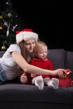 Anne ve küçük kızı yılbaşı hediyelerini evdeki koltukta, Noel ağacının arka planında açıyorlar. Dikey çerçeve.
