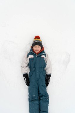 Sıcak tulumlu ve şapkalı çocuk karda yatıyor. Kış tatili Çocuk karda oyun oynuyor. Üst görünüm.