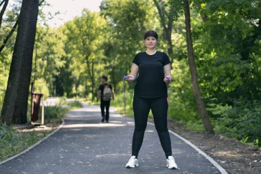 Ayrıca parkta ip atlayan bir kadın var ve kameraya bakıyor. Aktif giyimli genç bir kadın sağlıklı yaşam tarzını seçiyor. Vücut pozitifliği