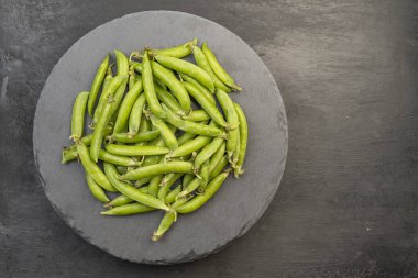 Siyah zemin üzerinde siyah yuvarlak bir tabakta olgun yeşil bezelye kabukları. Taze sebzeler. Sütunlar