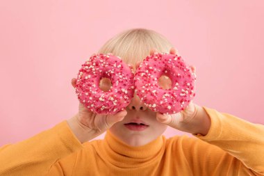 Fair-haired baby holds pink donuts near his eyes. Portrait of boy with doughnut on pink background