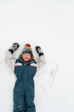 Grimacing boy with closed eyes in a warm jumpsuit and hat lies in the snow. Winter holidays. Child is playing in the snow. Top view.