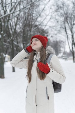 Brooding young woman in red knitted hat and mittens walks through snow-kept park. Winter day