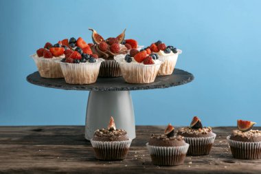 Confectionery on stand. Vanilla and chocolate muffins with fresh berries on blue background. Side view, copy space.