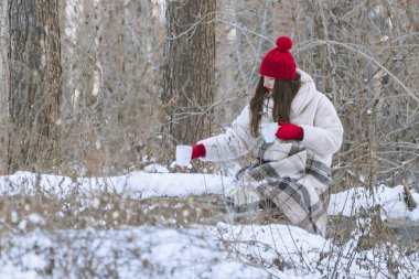 Brunette woman in red hat and mittens in winter forest with two mugs of coffee waiting for a friend.