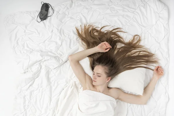 Beautiful young woman sleeps in her bed on white sheets at home. Top view portrait of sleeping girl with long loose hair