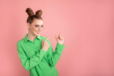 Young woman points away with fore finger, shows something. Girl wears glasses and green shirt. Stands over pink wall with space for your text