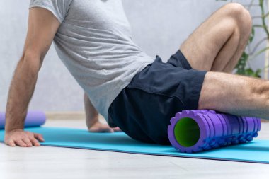 Man sits on yoga mat and doing massage his feet with foam roller. Workout at home. Roller for physiotherapy
