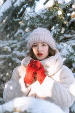 Portrait of beautiful girl in red mittens and big hat on snow-covered fir trees background. Vertical frame.