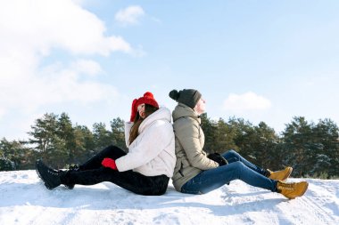 Genç bir adam ve kadın, karlı bir tepede arka planda, ormanın ve mavi gökyüzünün önünde sırt sırta oturuyorlar. Dışarıda bir çift..