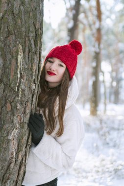 Kırmızı şapkalı güzel esmer kadın karlı bir parktaki bir ağaca sarılıp yatıyor. Açık havada portre. Dikey çerçeve.