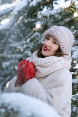 Güzel genç bir kadın, kar kaplı çam ağaçlarının arasında duruyor. Sıcak beyaz bir şapka ve kürk mantoyla. Güneşli kış günü. Dikey çerçeve. Boşluğu kopyala.