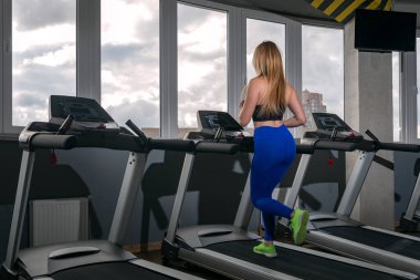 Spor kıyafetli sarışın bir kız modern spor salonunda koşu bandında koşuyor. Çalışan pistte kardiyo çalışması.