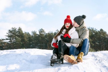 Gençler kış ormanı ve mavi gökyüzünün arka planında oturup sarılıyorlar. Kış güneşli bir gün. Boşluğu kopyala.