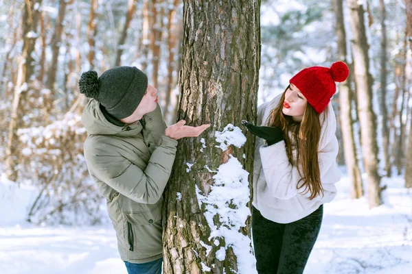 年轻夫妇在一棵树后窥视 并在雪地的森林里互相亲吻 在阳光灿烂的冬季公园里的男人和女人 — 图库照片