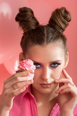Pembe ve mavi renkli parlak makyaj. İki çörekli, saç stili ve elinde kek olan güzel bir kızın portresi. Dikey çerçeve.