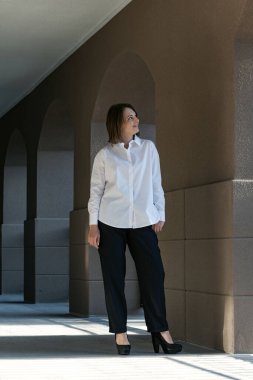 Beyaz gömlekli ve siyah pantolonlu gülümseyen genç kadın binanın dışında poz veriyor. Klasik kostümlü bir kızın boy portresi.