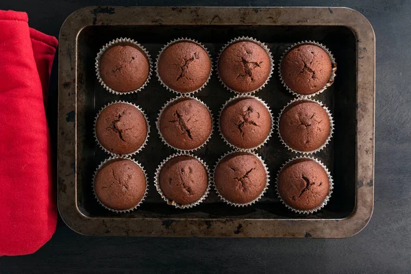 Chocolate brownies muffins on baking sheet and red pot-holders on black background. Just cooked homemade cupcake