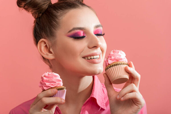 Девушка с профессиональным розовым макияжем наслаждается вкусные кексы. Портрет женщины с конфетами на розовом фоне