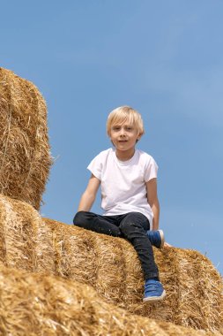 Mavi gökyüzü arka planında kuru saman yığınının üzerinde beyaz tişörtlü sarışın bir çocuğun portresi. Yaz tatili. Hasat.