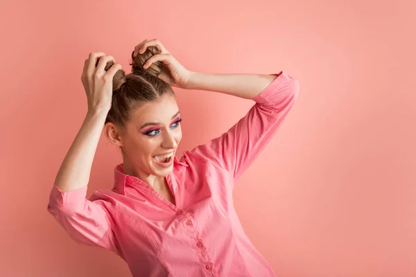 Funny young woman in pink blouse on pink background. Surprised laughing girl with two bun hairstyles looking away. Copy space.