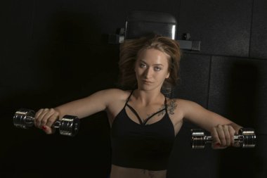 Atletik kadın sırt üstü uzanıyor ve spor salonunda antrenman yapıyor doğrudan kameraya bakıyor. Dambıl ile egzersiz. Üst görünüm.