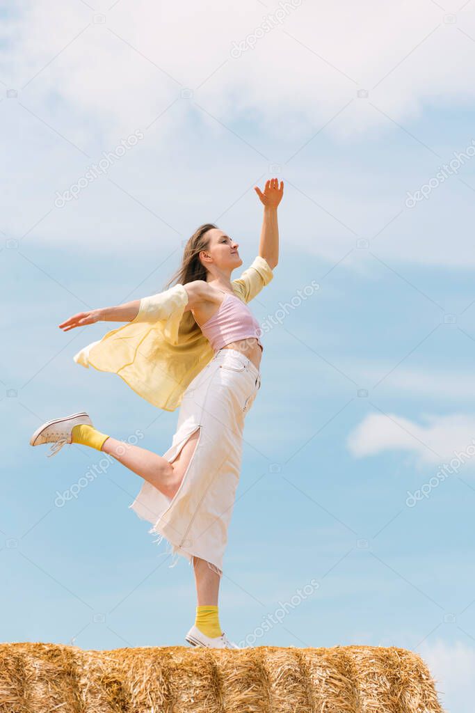 Portrait of young attractive dancing woman on top of haystack on blue sky background. Vertical frame