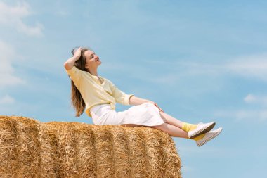 Genç bir kadın büyük saman yığınlarının üzerinde oturuyor. Samanlıkta hasattan sonra tarlada poz veren kadın model.