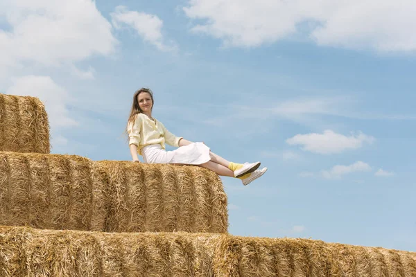 Yaz tarlasında saman balyasında dinlenen bir kız. Mavi gökyüzü arka planında samanlıkta oturan genç ve güzel bir kadın.
