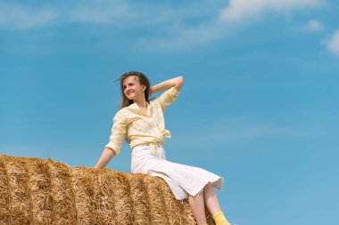 Samanlıkta oturan güzel genç bir kadın, mavi arka planda kırsal kesimde. Yüksek samanlıkta bir kızın portresi