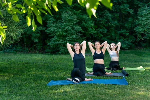Üç kız yoga minderlerine oturup esneme hareketleri yapıyor. Parkta grup yoga çalışması. Sabah sporu.
