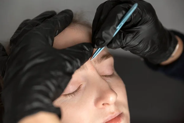 Procedure of eyebrow correction, young woman in beauty salon. Cosmetologist in black gloves plucks eyebrows with tweezers