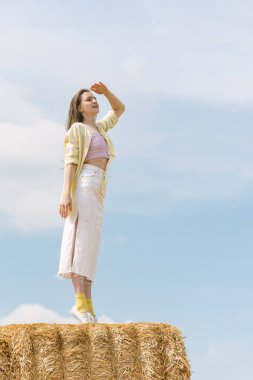 Güzel genç bir kadın yüksek samanlıkta duruyor ve uzaklığa bakıyor. Kırsal tatil. Saman hasatı.