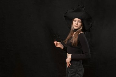 Cadı kılığındaki genç kadın siyah elbiseler giyiyor, şapka takıyor ve gözlük tutuyor. Uzayı kopyala Siyah arkaplanda izolasyon.