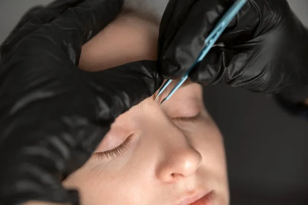 Portrait of young woman on the procedure of eyebrow correction. Cosmetologist in black gloves plucks eyebrows with tweezers