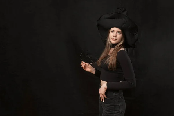 Young woman in form of witch wears black clothes, pointed hat and holds glasses. Copy space. Isolation on black background.