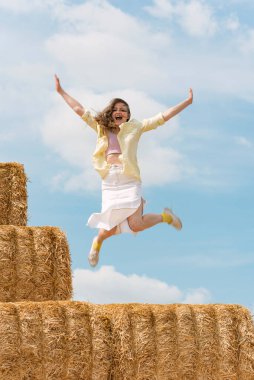 Neşeli kız samanlıktan atlıyor. Mutlu genç kadın mavi gökyüzü arka planında çiftlikte eğleniyor.