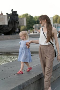 Genç anne küçük kızıyla meydanda yürüyor. Mavi elbiseli küçük kız annesini ya da bakıcısını elinde tutuyor.