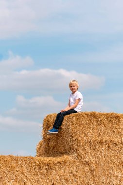 Samanlıkta oturan sarışın çocuğun dikey portresi. Çocuk köyde tatilde.