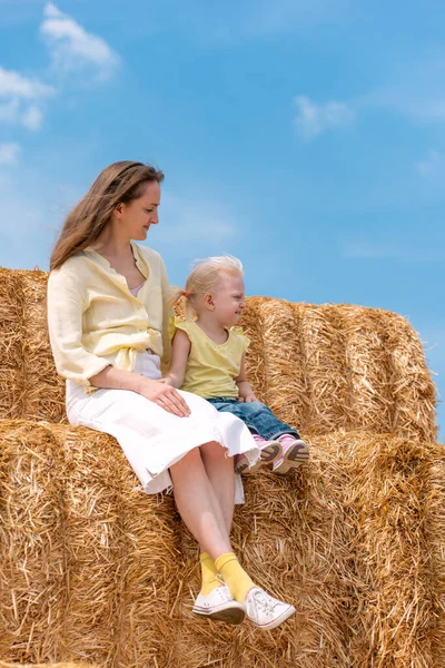 Mother with little happy daughter on huge haystack on background of blue sky. Vertical frame. Mom and fair-haired girl are sitting in the hay