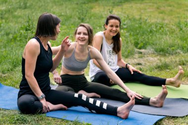 Üç genç gülen kız çimlerin üzerinde spor yapıyor. Atletik kadınlar kalçalarının arkasına uzanıyor..