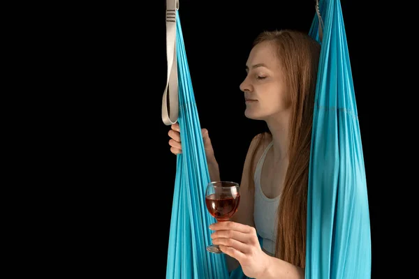 Young woman sits in gymnastic hammock with glass of wine in her hands. Meditation, sports and alcohol