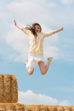Gülen kız samanlıktan atlıyor. Mutlu genç kadın mavi gökyüzü arka planında çiftlikte eğleniyor.