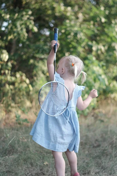 金发3岁的女孩 有两个尾巴和夏美裙 打羽毛球 穿蓝色衣服的孩子拿着球拍和羽毛球 — 图库照片